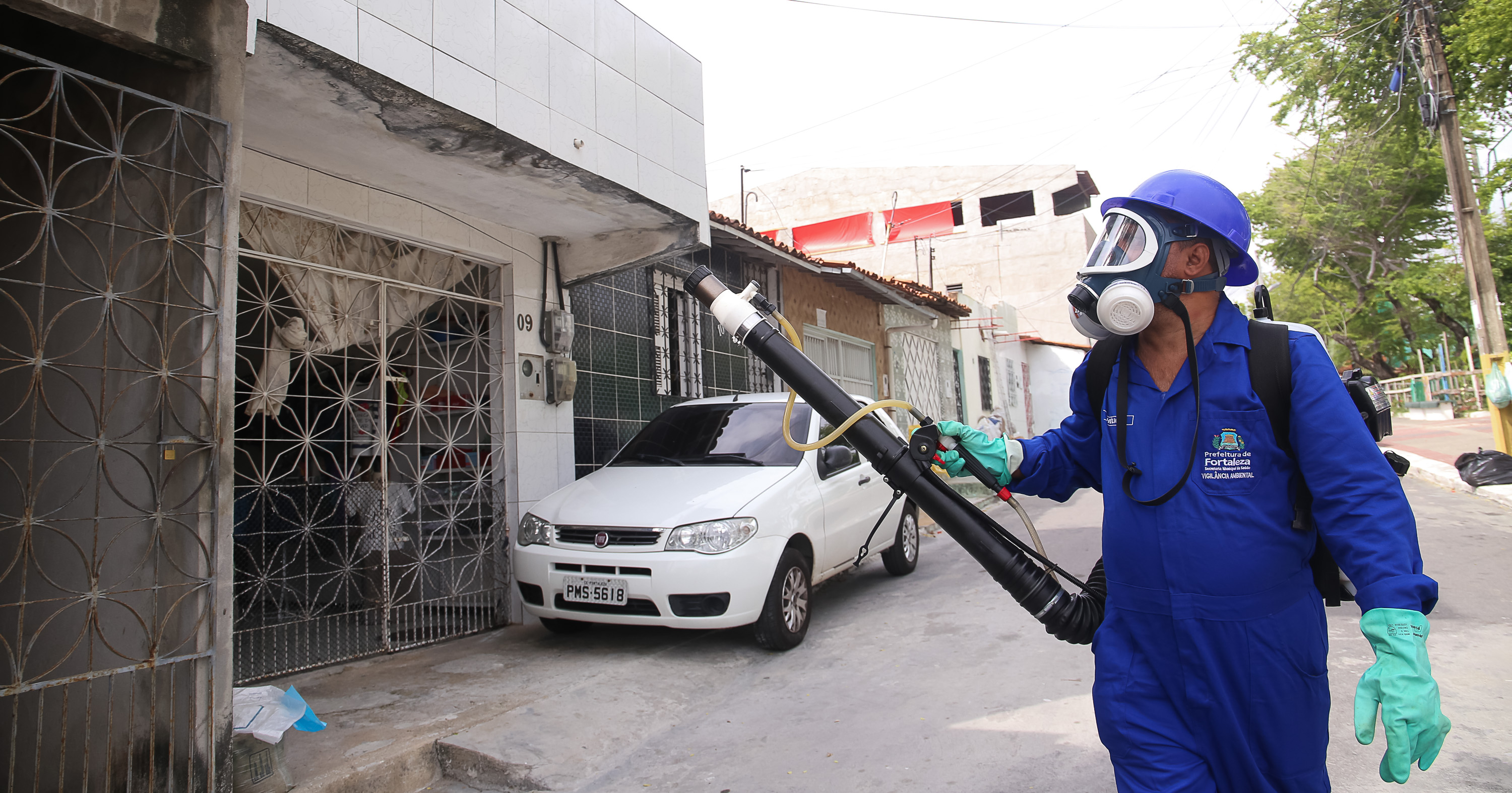 homem, como equipamento de proteção, ou seja, macacão e máscara completa no rosto, passa com a mochila do fumacê e a mangueria que solta a fumaça. Ele caminha por uma rua no Vicente Pinzón